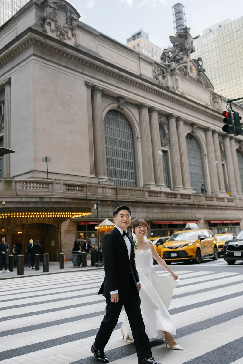 The couple walking together across a city crosswalk in front of a grand station.