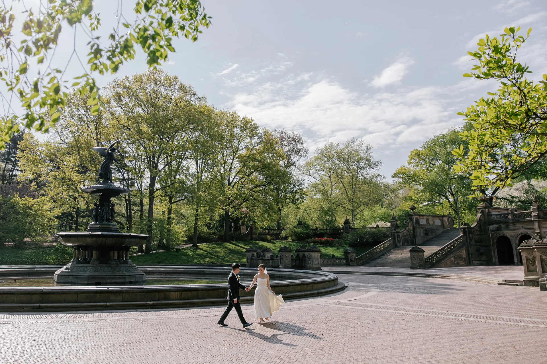 The couple walking hand in hand in front of a fountain, surrounded by spring greenery.